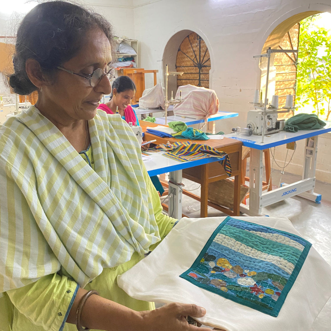 Woman holding a quilt with a colorful design in a room with various items and people in the background.