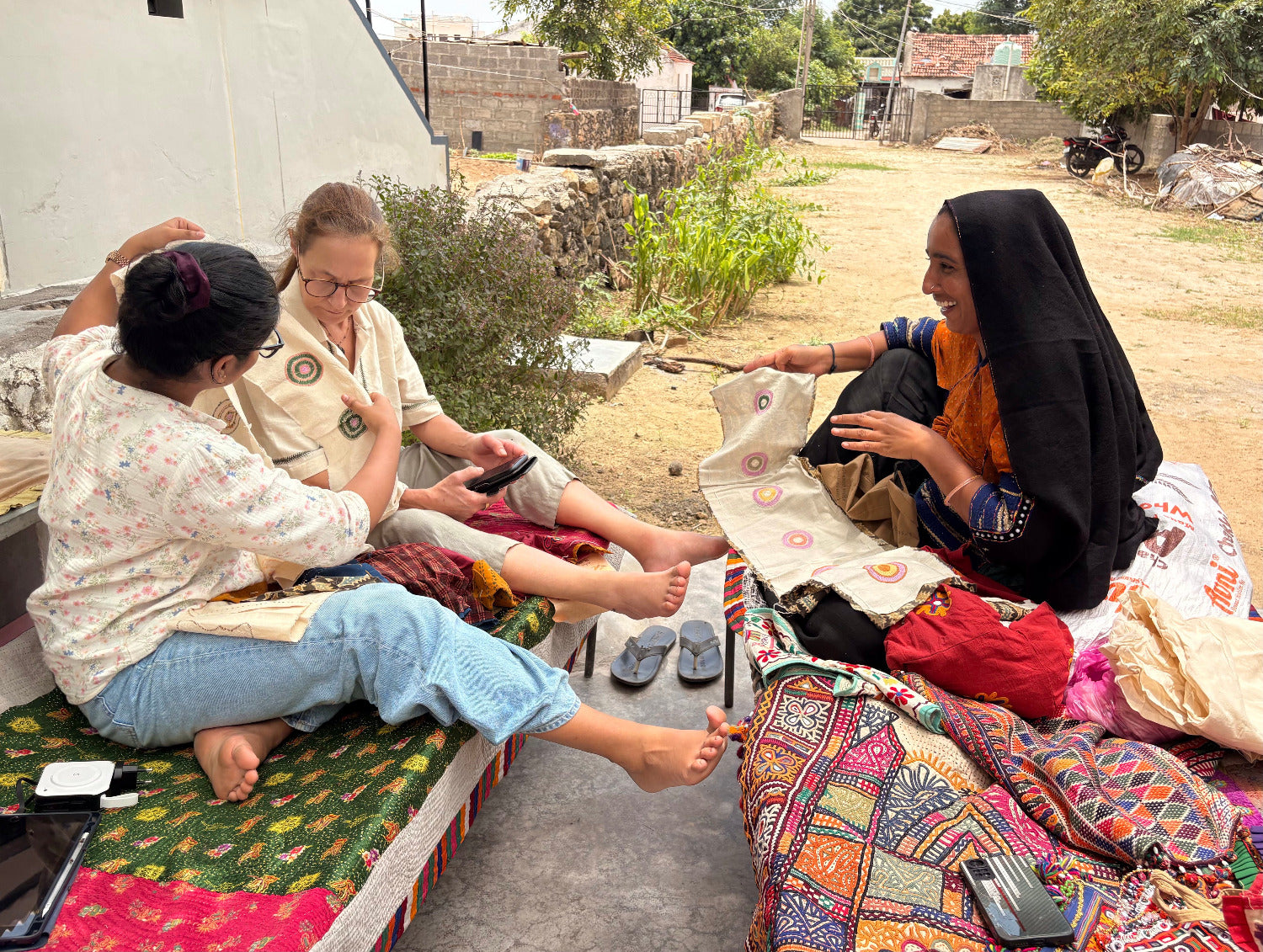Three women sitting on a bench in an outdoor setting with colorful fabrics and shoes.