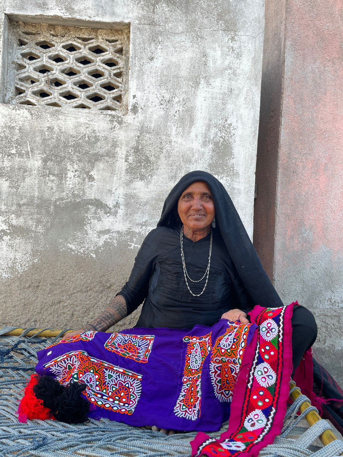 Woman sitting on a bed with a colorful blanket against a plain wall.