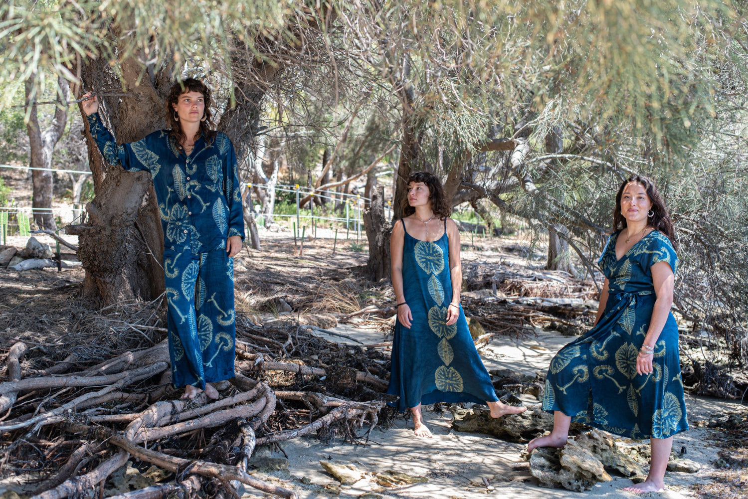 Three women in blue dresses standing among trees and fallen branches.